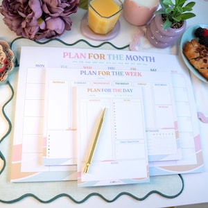 May include: A set of pastel-colored planning pads with the text "Plan for the Month", "Plan for the Week", and "Plan for the Day". A gold pen rests on the smallest pad. The pads have a wavy design.