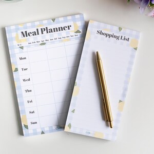 Op de afbeelding: Een set van twee notitieblokken met een blauw-wit geruit patroon en citroenillustraties. Het bovenste notitieblok is gelabeld "Meal Planner" en heeft een wekelijks kalenderformaat. Het onderste notitieblok is gelabeld "Shopping List".