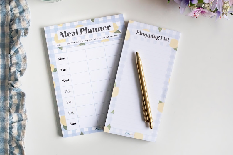 Op de afbeelding: Een set van twee notitieblokken met een blauw-wit geruit patroon en citroenillustraties. Het bovenste notitieblok is gelabeld "Meal Planner" en heeft een wekelijks kalenderformaat. Het onderste notitieblok is gelabeld "Shopping List".