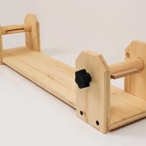 Peut inclure: Un dévidoir à fil en bois clair, conçu pour maintenir la laine lors de l'enroulement. Il est composé d'une base rectangulaire et de deux supports verticaux, chacun avec une cheville en bois et un bouton noir pour le réglage.