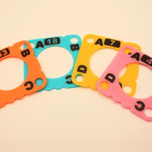 May include: Four colorful, square-shaped foam pieces with a circular cutout in the center. Each piece is a different color: orange, teal, yellow, and pink. Black letters and numbers are printed on each piece, including A33, A18, A7, and A4.