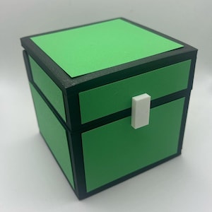 May include: A green and black square box with a white rectangular clasp. The box has a green top and green sides with black trim. The box is a cube shape.