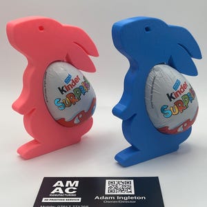 May include: Two 3D printed bunny rabbit holders, one pink and one blue, holding a Kinder Surprise egg. The business card of AM AC Manufacturing, a 3D printing service, is in the foreground. The card includes the company name, contact information, and the text "Design & Supply 3D Printing Services".