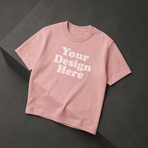 May include: A light pink t-shirt with short sleeves and the text "Your Design Here" in white. The shirt has a crew neck and is made of a soft material. It is displayed on a dark grey surface.