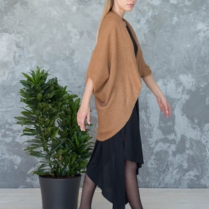 May include: A woman wearing a brown knitted cardigan over a black asymmetrical dress and black tights. She is standing in front of a gray wall with a potted plant.