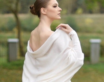 May include: A white knitted shawl draped over a person's shoulders. The shawl has a cowl neck and long sleeves. The person is wearing a white dress.
