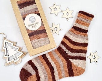 Könnte beinhalten: Eine gestreifte Socke in Braun-, Beige- und Cremetönen, neben einer passenden Socke in einer Schachtel. Auf der Schachtel befindet sich ein Etikett mit der Aufschrift "SANDRA MG". Holzsterne und -baumschmuck sind um die Socke herum angeordnet, was auf ein festliches Thema hindeutet.