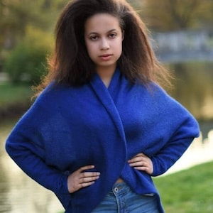 May include: A young woman with long, dark hair is wearing a blue sweater and blue jeans. She is standing in a park with green grass and a body of water in the background.