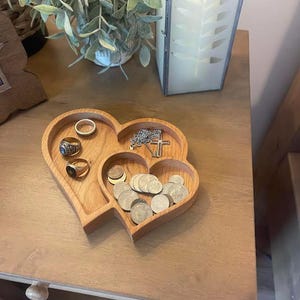 Op de afbeelding: Een houten hartvormig sieradenbakje met drie compartimenten. Het bakje bevat ringen, munten en een ketting met een kruisvormige hanger. Het bakje is gemaakt van lichtgekleurd hout.