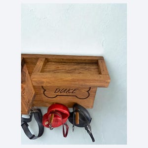 Pu&ograve; includere: Porta guinzaglio e dolcetti per cani in legno con il nome "DUKE" intagliato sul davanti. Il supporto ha un design a forma di osso e include un ripiano. Sotto il supporto ci sono un guinzaglio nero, un contenitore rosso per dolcetti e una borsa nera.