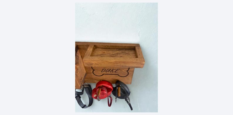 Pu&ograve; includere: Porta guinzaglio e dolcetti per cani in legno con il nome "DUKE" intagliato sul davanti. Il supporto ha un design a forma di osso e include un ripiano. Sotto il supporto ci sono un guinzaglio nero, un contenitore rosso per dolcetti e una borsa nera.