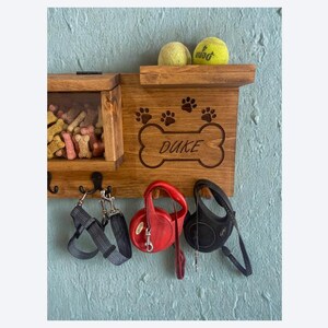 Pu&ograve; includere: Organizzatore per guinzagli e snack per cani in legno con il nome "Duke" su un design a forma di osso. Include un ripiano con due palline da tennis, un vano per snack e ganci per guinzagli e imbracature. Montato su una parete blu.