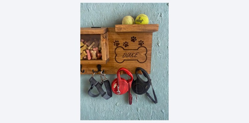 Pu&ograve; includere: Organizzatore per guinzagli e snack per cani in legno con il nome "Duke" su un design a forma di osso. Include un ripiano con due palline da tennis, un vano per snack e ganci per guinzagli e imbracature. Montato su una parete blu.