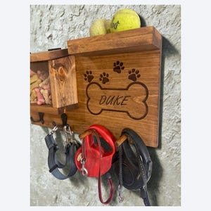 Pu&ograve; includere: Organizzatore in legno per guinzagli e snack per cani con il nome "Duke" inciso su un design a forma di osso. Presenta impronte di zampe, ganci per guinzagli e un ripiano con palline da tennis. Montato su una parete strutturata.