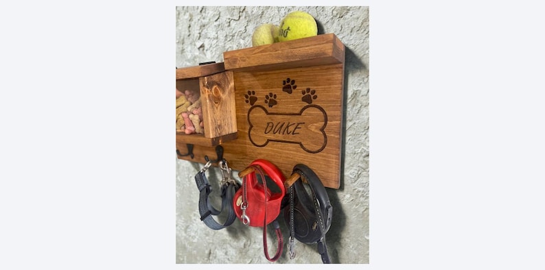 Pu&ograve; includere: Organizzatore in legno per guinzagli e snack per cani con il nome "Duke" inciso su un design a forma di osso. Presenta impronte di zampe, ganci per guinzagli e un ripiano con palline da tennis. Montato su una parete strutturata.