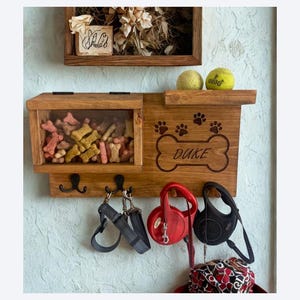 Pu&ograve; includere: Organizzatore in legno per snack e guinzagli per cani con un vano anteriore trasparente pieno di snack per cani. L'organizzatore ha ganci per guinzagli, un ripiano con due palline da tennis e un design a forma di osso con il nome "Duke".