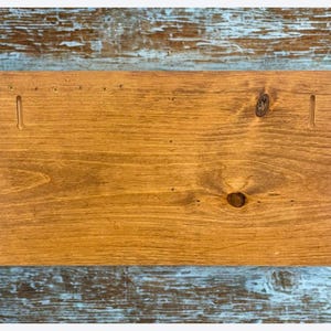 Pu&ograve; includere: Un cartello rettangolare in legno con una calda macchia marrone dorata. La venatura del legno &egrave; visibile e ci sono due fessure intagliate vicino alla parte superiore per appenderlo. Il cartello &egrave; su uno sfondo blu e bianco usurato.
