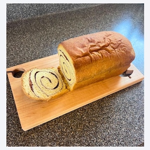 May include: A loaf of cinnamon swirl bread, partially sliced, rests on a light-colored wooden cutting board. The bread has a golden-brown crust and a visible swirl pattern. The cutting board has a handle-like cutout on one side.