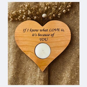 May include: A heart-shaped wooden candle holder with the inscription "If I know what LOVE is, it's because of YOU." A white candle sits in the center. The wood has a warm, natural tone. Decorative sprigs are at the top.