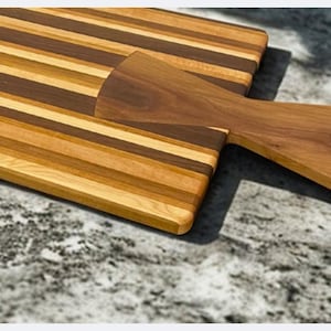 May include: Wooden cutting board set with a striped design. The set includes a rectangular board and a paddle-shaped board with a handle. The boards are made of various shades of brown wood.