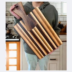 May include: A large wooden cutting board with a handle, featuring alternating stripes of light and dark wood. The board is rectangular, with a handle on one end. The cutting board is approximately 18 inches long and 12 inches wide.