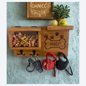 Pu&ograve; includere: Organizzatore in legno per cani con il nome "Duke" su un design a forma di osso. Dispone di un vano trasparente per i dolcetti, ganci per guinzagli e un ripiano con una piccola pianta e palline da tennis. Sfondo blu strutturato.