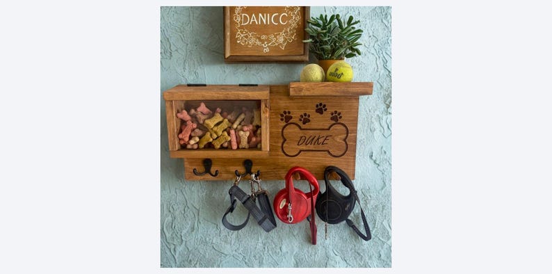 Pu&ograve; includere: Organizzatore in legno per cani con il nome "Duke" su un design a forma di osso. Dispone di un vano trasparente per i dolcetti, ganci per guinzagli e un ripiano con una piccola pianta e palline da tennis. Sfondo blu strutturato.