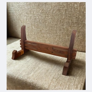 Op de afbeelding: Een houten displaystandaard met een rijke bruine afwerking. De standaard heeft een horizontale basis, twee verticale palen en twee schuine steunen. Het ontwerp is eenvoudig en functioneel, geschikt voor het presenteren van diverse items. Ongeveer 25 cm lang.