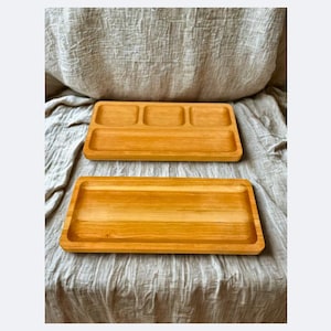 May include: Two rectangular wooden serving trays. The top tray has three compartments, while the bottom tray is a single, open space. Both trays are made of light-coloured wood and are set against a neutral-toned fabric background.