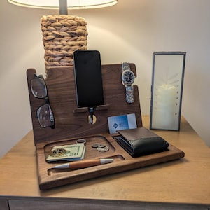 May include: A wooden bedside organizer with compartments for a phone, glasses, watch, wallet, cash, and pen. The organizer is made of dark wood and sits on a wooden nightstand. A lamp with a woven base is in the background.