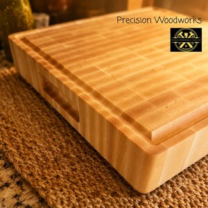 K&ouml;nnte beinhalten: Ein dickes, helles Stirnholz-Schneidebrett mit glatter Oberfl&auml;che, einer umlaufenden Saftrille und integrierten Seitengriffen. Der Text Precision Woodworks sowie ein kreisf&ouml;rmiges Logo sind in der Ecke des Brettes deutlich zu erkennen.