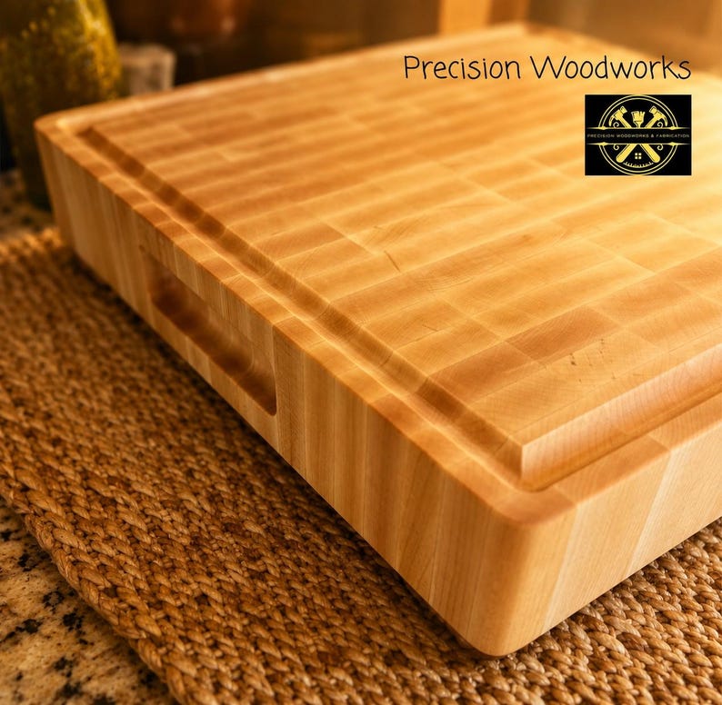 K&ouml;nnte beinhalten: Ein dickes, helles Stirnholz-Schneidebrett mit glatter Oberfl&auml;che, einer umlaufenden Saftrille und integrierten Seitengriffen. Der Text Precision Woodworks sowie ein kreisf&ouml;rmiges Logo sind in der Ecke des Brettes deutlich zu erkennen.