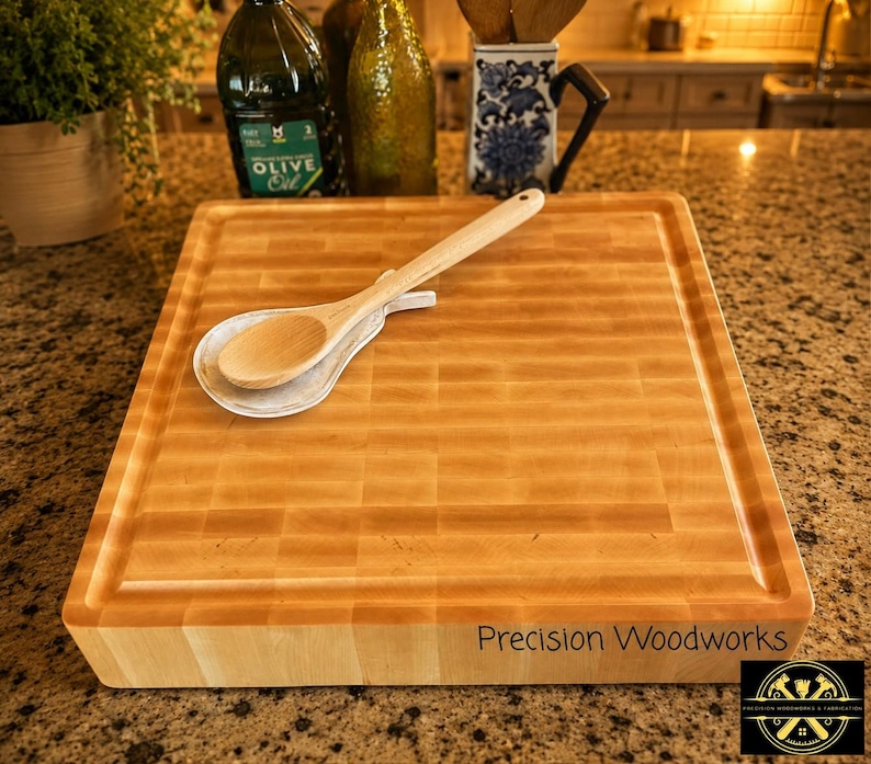 K&ouml;nnte beinhalten: Ein rechteckiges, helles Holzschneidebrett mit einem gitterartigen Hirnholzmuster und einer umlaufenden Saftrille. Der Text Precision Woodworks ist auf der Vorderkante eingraviert. Ein wei&szlig;er Keramik-L&ouml;ffelablage und ein Holzl&ouml;ffel liegen darauf.