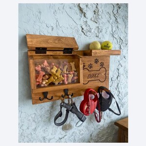 Pu&ograve; includere: Organizzatore in legno per snack e guinzagli per cani con coperchio incernierato, parte anteriore trasparente e ganci. L'organizzatore ha il nome "Duke", insieme a impronte di zampe e un disegno a forma di osso. Due palline da tennis sono su una mensola.