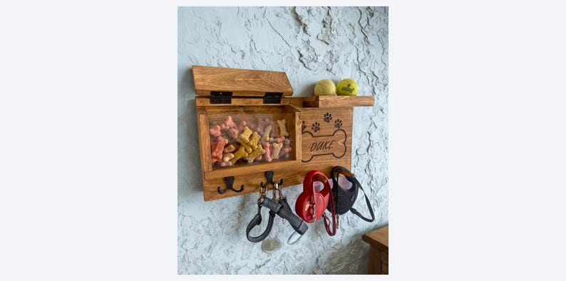 Pu&ograve; includere: Organizzatore in legno per snack e guinzagli per cani con coperchio incernierato, parte anteriore trasparente e ganci. L'organizzatore ha il nome "Duke", insieme a impronte di zampe e un disegno a forma di osso. Due palline da tennis sono su una mensola.