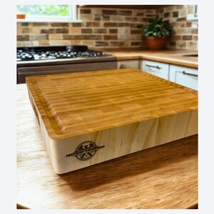 K&ouml;nnte beinhalten: Ein dickes, quadratisches Hackblock-Schneidebrett aus Holz mit einer Stirnholzoberfl&auml;che. Die Seitenwand aus hellem Holz ist mit einem kreisf&ouml;rmigen schwarzen Logostempel versehen, der "PREMIUM WOODWORKS" und ein stilisiertes Bergsymbol zeigt.
