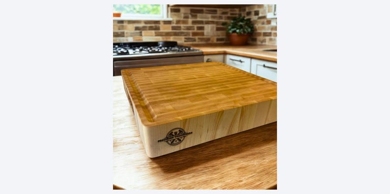 K&ouml;nnte beinhalten: Ein dickes, quadratisches Hackblock-Schneidebrett aus Holz mit einer Stirnholzoberfl&auml;che. Die Seitenwand aus hellem Holz ist mit einem kreisf&ouml;rmigen schwarzen Logostempel versehen, der "PREMIUM WOODWORKS" und ein stilisiertes Bergsymbol zeigt.