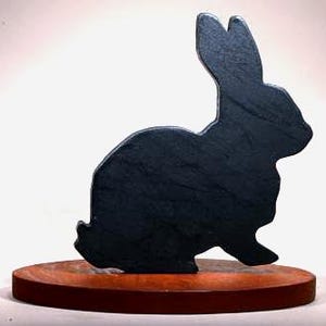 May include: A dark gray rabbit silhouette sculpture on a round, reddish-brown wooden base. The rabbit is in a seated position with its ears upright. The sculpture is likely a decorative item.