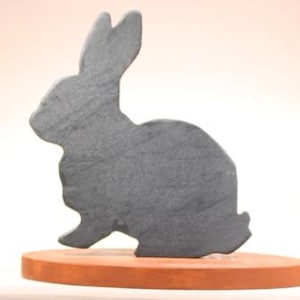 May include: A dark gray, flat, rabbit-shaped silhouette cutout made of stone or slate material, mounted vertically onto a smooth, oval-shaped, terracotta-colored base. The rabbit figure is depicted in profile facing left with upright ears.