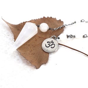 May include: A white, translucent, faceted crystal pendulum attached to a silver-toned metal chain. The chain features a round, silver-toned metal charm embossed with the Om symbol and a single, smooth, round white bead near the crystal point.