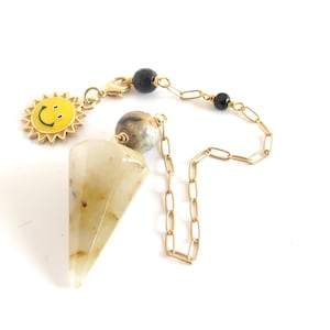 May include: A pendulum with a gold chain and a triangular, light-colored crystal. A yellow sun charm with a smiling face hangs from the chain. Black beads and a patterned bead are also attached.