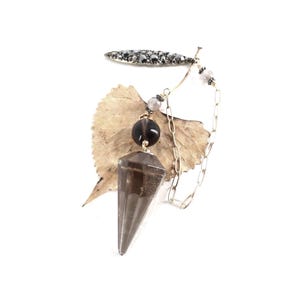 May include: A dowsing pendulum featuring a faceted, translucent smoky quartz point suspended from a delicate gold-tone chain. The chain includes a dark, round bead, a smaller light-colored bead, and a decorative, elongated, dark-studded metal bar.