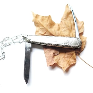 May include: A silver pocket knife with an ornate handle and a partially opened blade, attached to a silver chain. The knife rests on a dried, brown maple leaf against a white background.