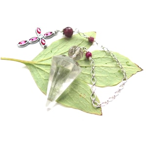 May include: A silver-toned necklace featuring a cross pendant with pink accents and a clear crystal point. The necklace includes red gemstone beads and a chain, resting on a green leaf against a white background.