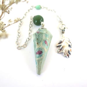 May include: A pendulum with a faceted, light green stone with red inclusions. It hangs from a silver chain with a green bead and a silver leaf charm. The background is white.