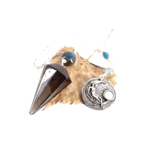 May include: A dark brown, faceted, cone-shaped pendulum attached to a silver chain, paired with a round, silver-toned metal pendant featuring a relief of a figure sitting on a crescent moon, accented with small blue and translucent beads.