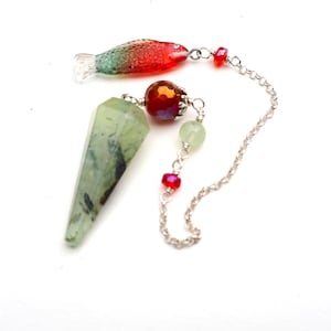 May include: A pendulum featuring a faceted, light green stone with dark inclusions, attached to a silver-toned chain. The chain includes a red and green glass fish charm, a large faceted red bead, a small pale green bead, and a tiny red bead.