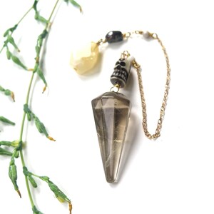 May include: A pendulum with a faceted, smoky quartz crystal point. The pendulum features a small skull bead, a gold-colored chain, and a cluster of beads. A sprig of green foliage is in the background.