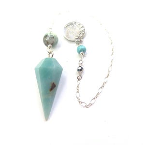 May include: A pendulum with a faceted, light blue gemstone. The pendulum is suspended by a silver chain and features decorative beads and a circular silver accent. The gemstone has a brown inclusion.