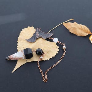 May include: A pendulum featuring a pointed black and white stone, a round black bead, and a copper-colored metal bat charm. The components are connected by a copper-colored chain with small white and metallic beads, resting on a dried leaf.